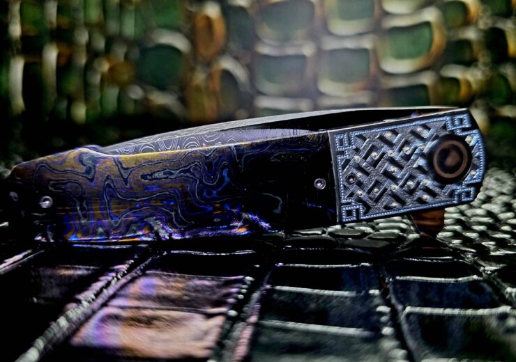 Herucus Blomerus custom folding knife open on dark surface showing carbon fiber handle and satin drop point blade