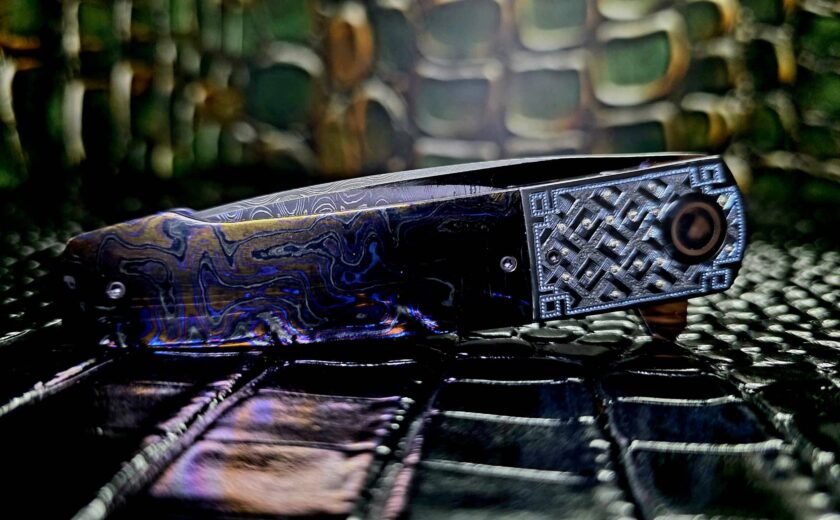 Herucus Blomerus custom folding knife open on dark surface showing carbon fiber handle and satin drop point blade