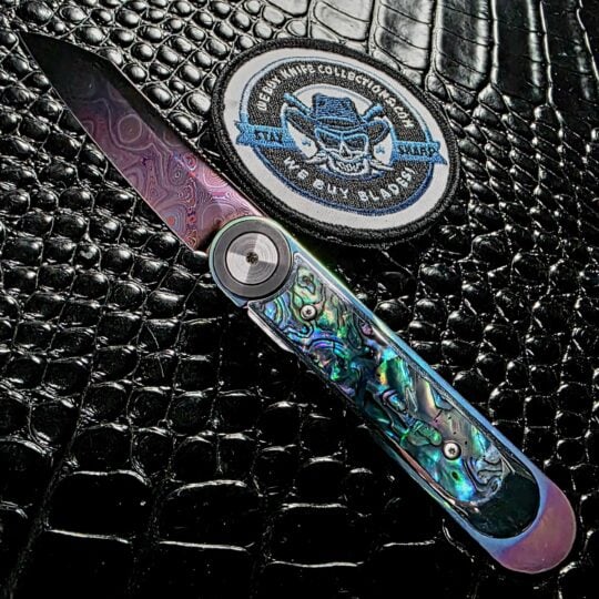 Dew Hara Infinity custom front flipper 1 of 1 open on the show side beside the We Buy Knife Collections patch, abalone scales and blued pyramid damascus blade
