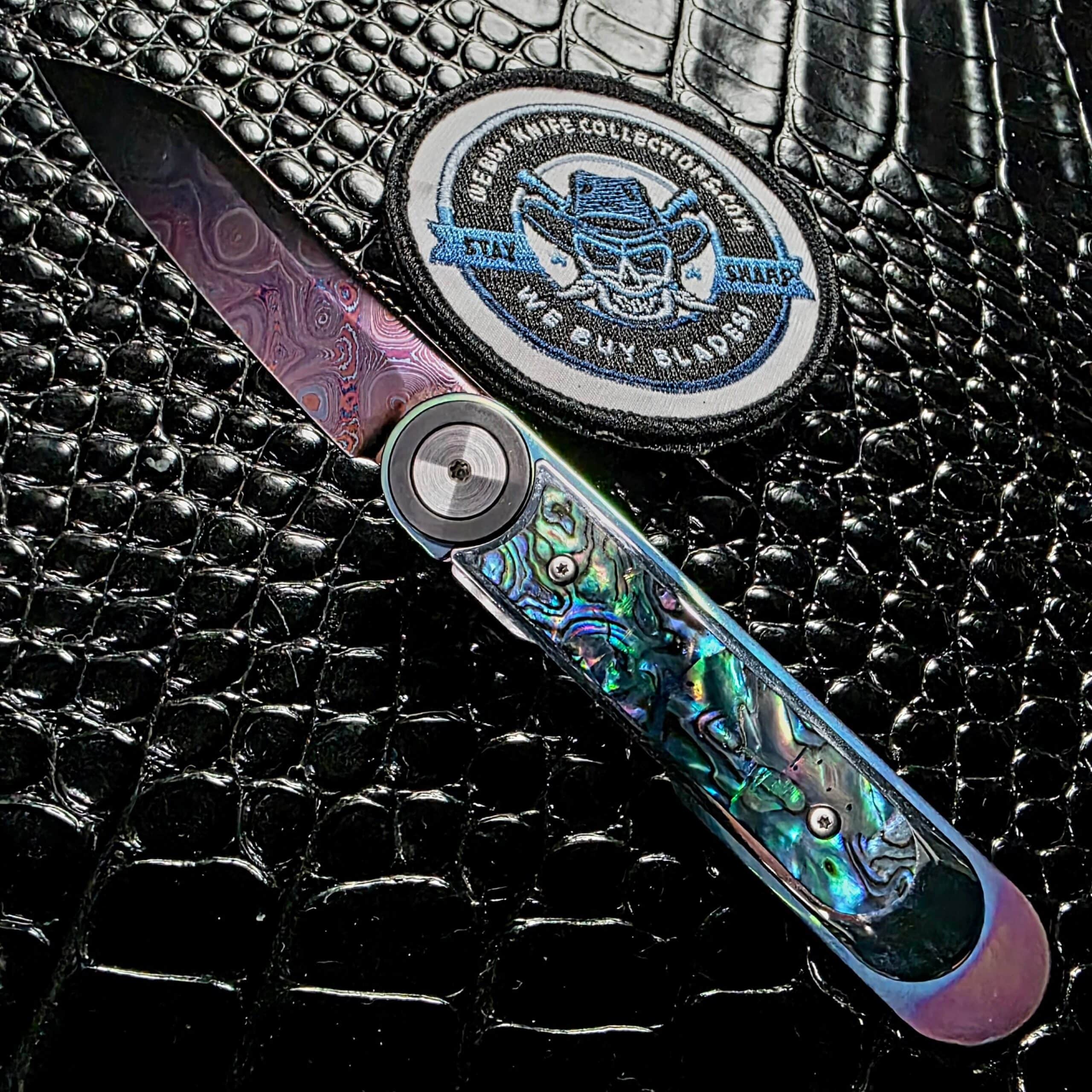 Dew Hara Infinity custom front flipper 1 of 1 open on the show side beside the We Buy Knife Collections patch, abalone scales and blued pyramid damascus blade