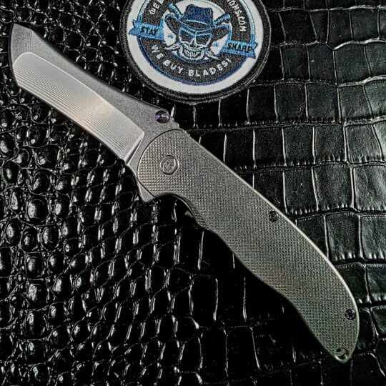 Grimsmo Norseman 5683 open on the show side beside the We Buy Knife Collections patch, silver Crosshatch titanium and stonewashed RWL 34 blade