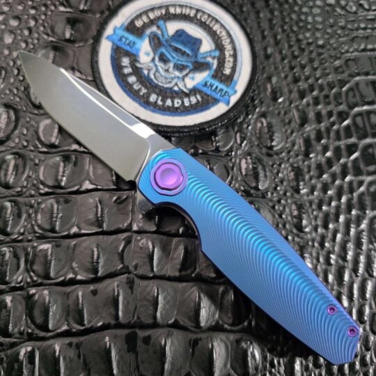 Holt Bladeworks Morpheus #1856 open on black crocodile leather with Near Mirror Vanax blade, Triad-milled blue titanium, and purple Black Timascus pivot collar