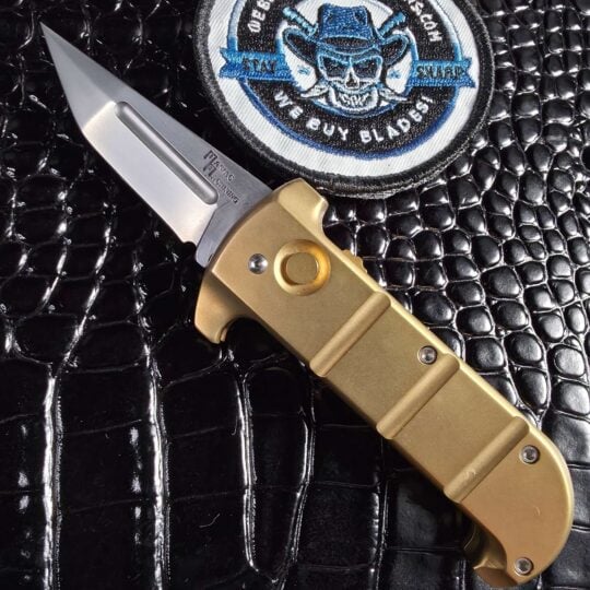 Maniac Machining Stinger EDC in solid 360 brass open beside the We Buy Knife Collections patch