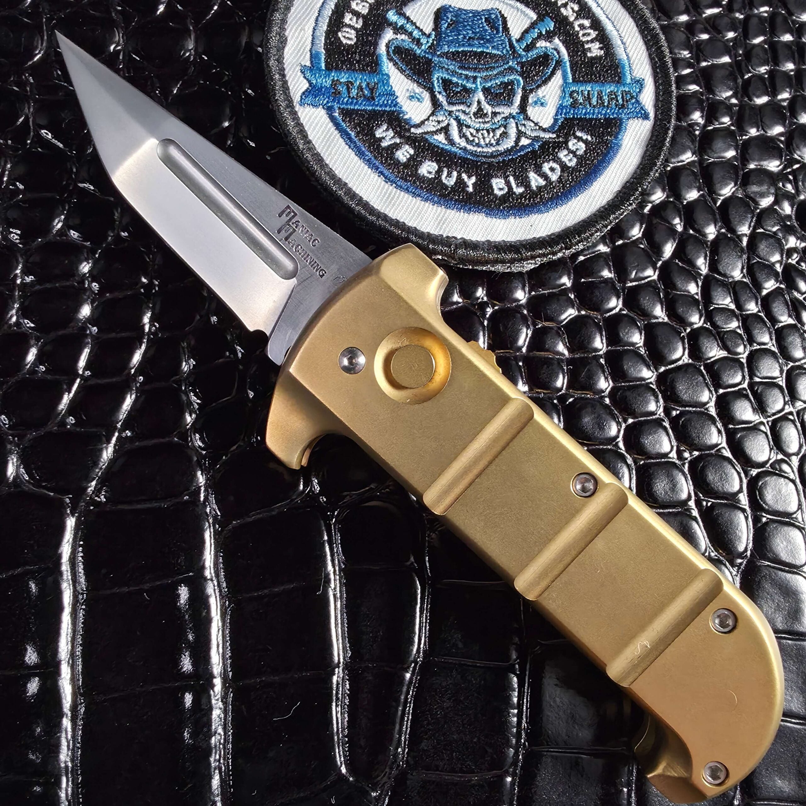 Maniac Machining Stinger EDC in solid 360 brass open beside the We Buy Knife Collections patch