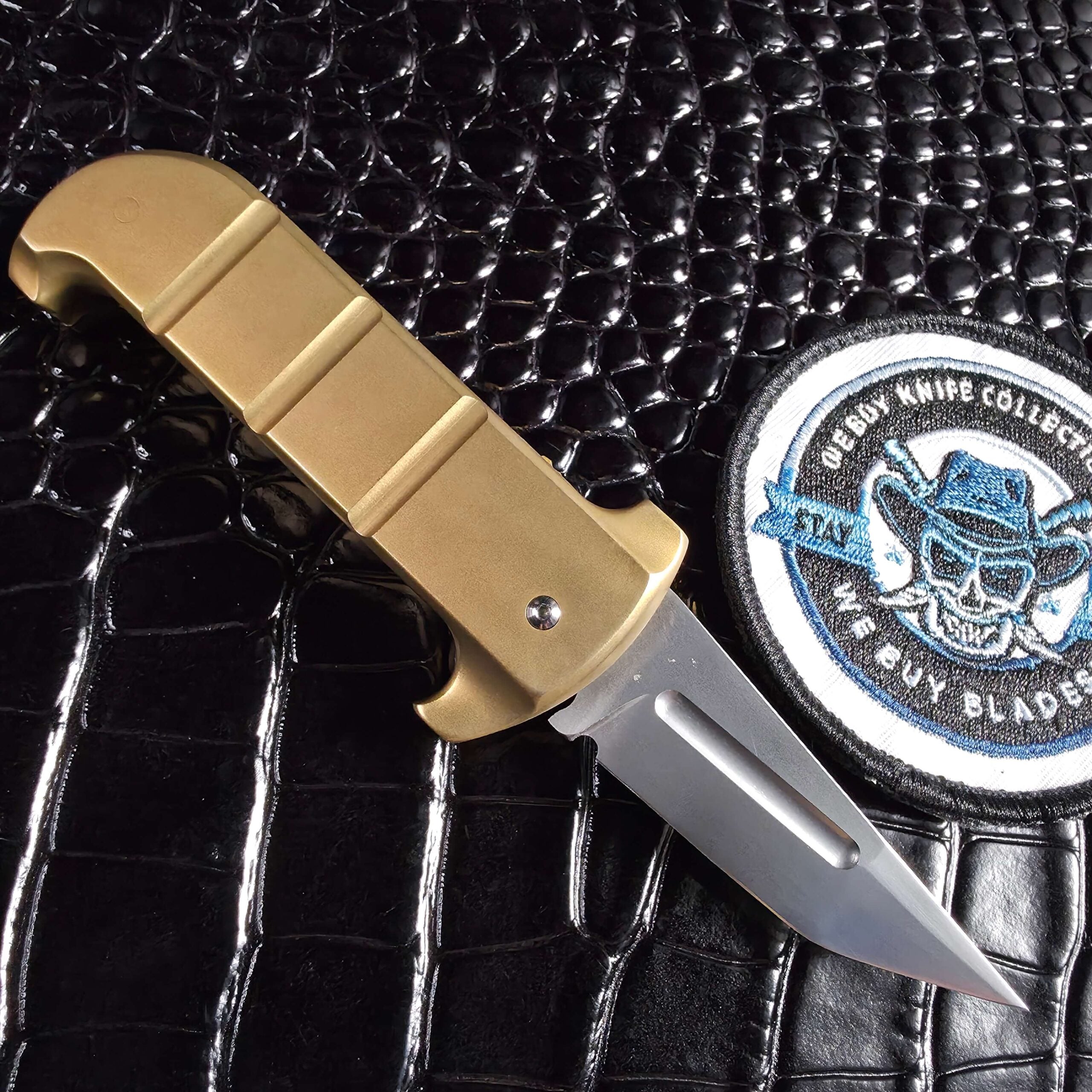 Reverse side of the Maniac Machining Stinger EDC open beside the We Buy Knife Collections patch