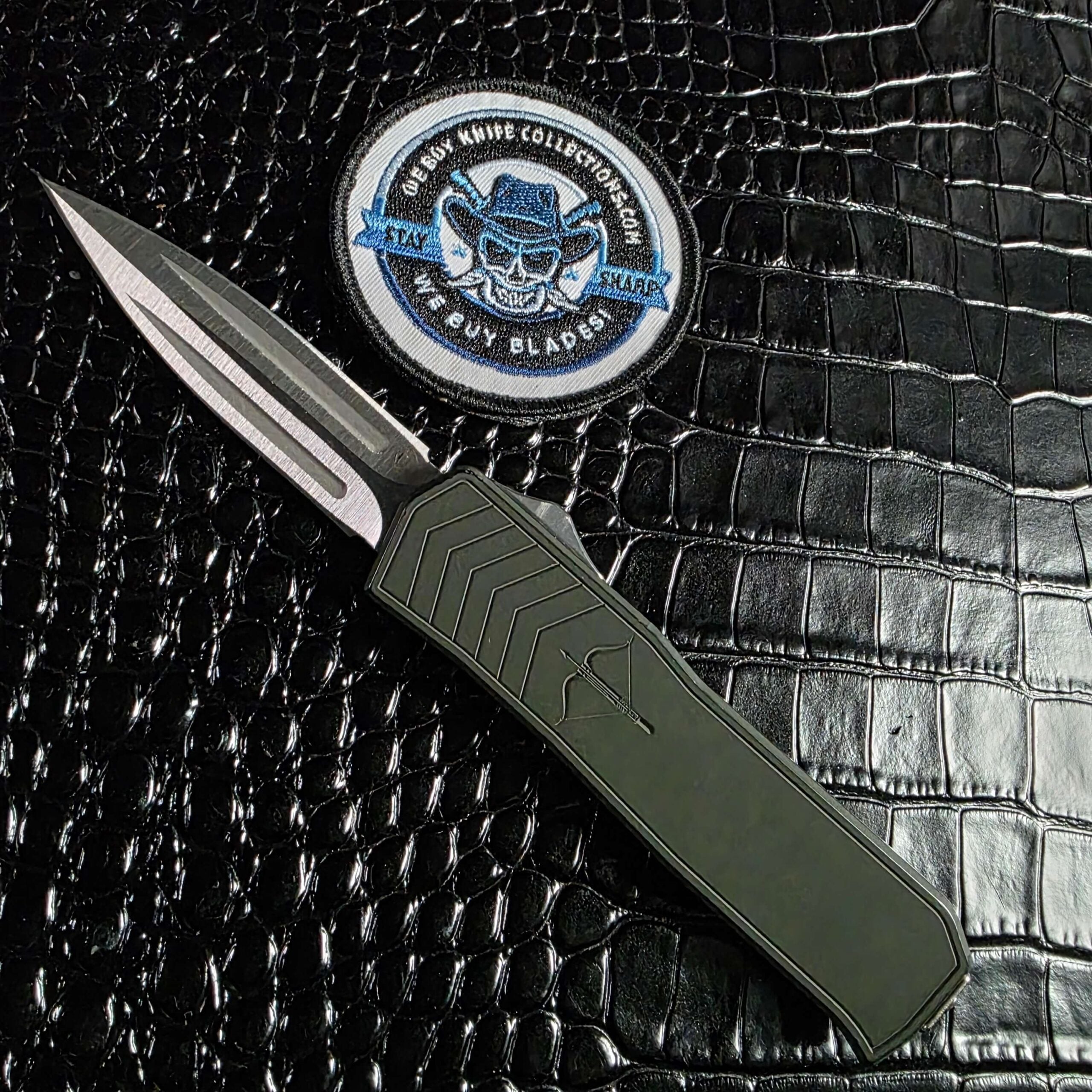 Medford Merc-1 OTF automatic dagger open beside the We Buy Knife Collections patch, 154CM blade and green aluminum handle