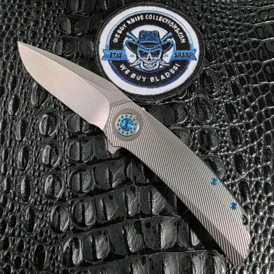 Skiff Made Blades Fugitive #0113 open on black crocodile leather with stonewashed Elmax blade, milled titanium scales, and seafoam anodized pivot collar