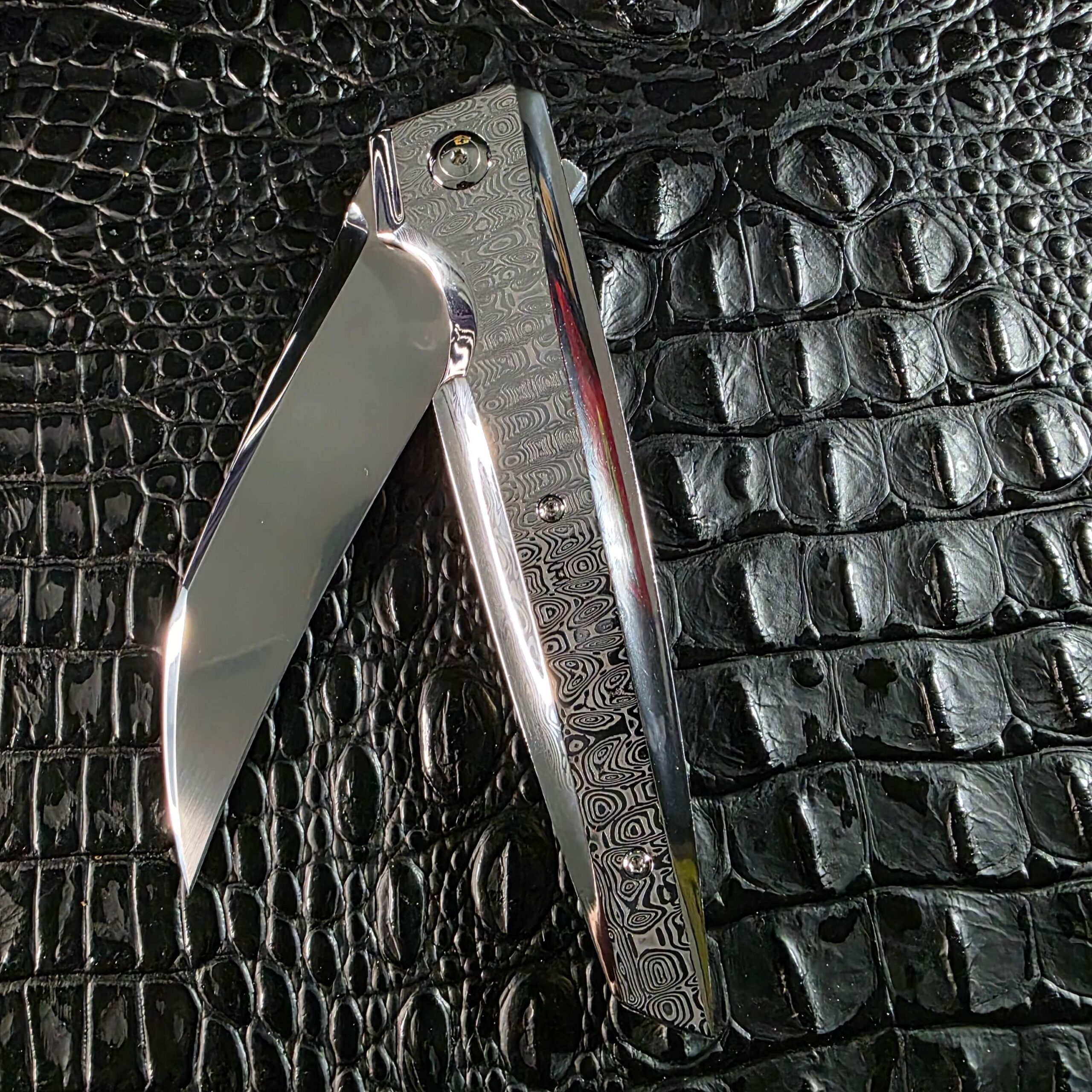 Stefano Compostella StriaXL shown half open on black crocodile leather, revealing the titanium liner lock and Damasteel Ladder scale
