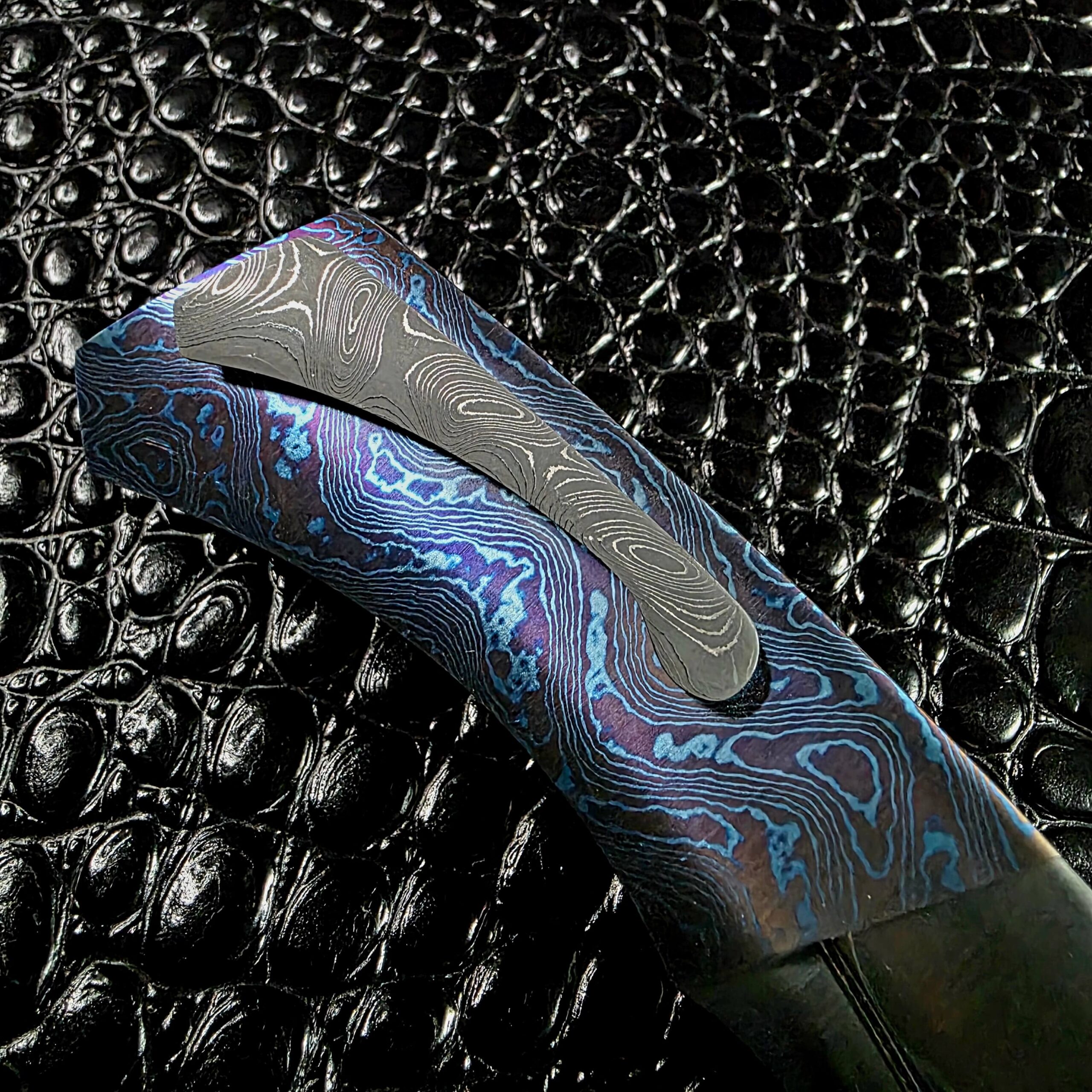 San Mai damascus pocket clip lying across the mokuti scale of the Kamehameha