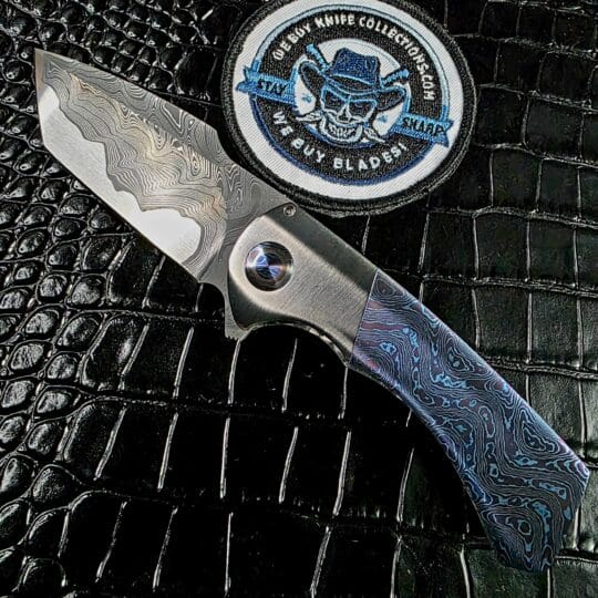 Todd Fischer Jr. Kamehameha custom flipper open on the show side beside the We Buy Knife Collections patch, mokuti scales and San Mai damascus blade