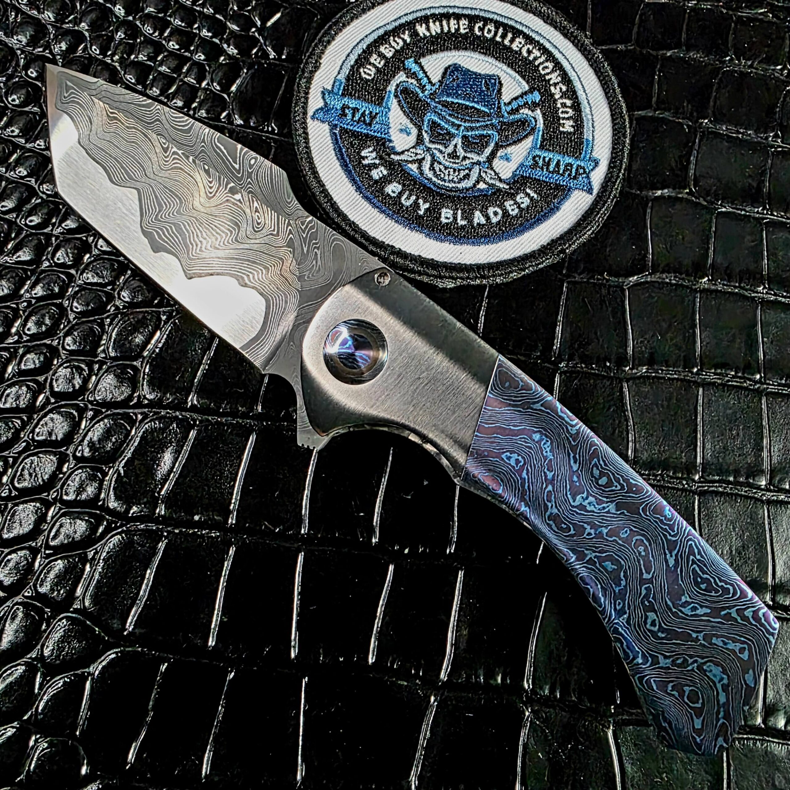 Todd Fischer Jr. Kamehameha custom flipper open on the show side beside the We Buy Knife Collections patch, mokuti scales and San Mai damascus blade