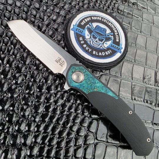 3LegDog M10 open on the show side, hand-rubbed satin AEB-L drop point above a teal stabilised burl inlay, with the We Buy Knife Collections patch in frame