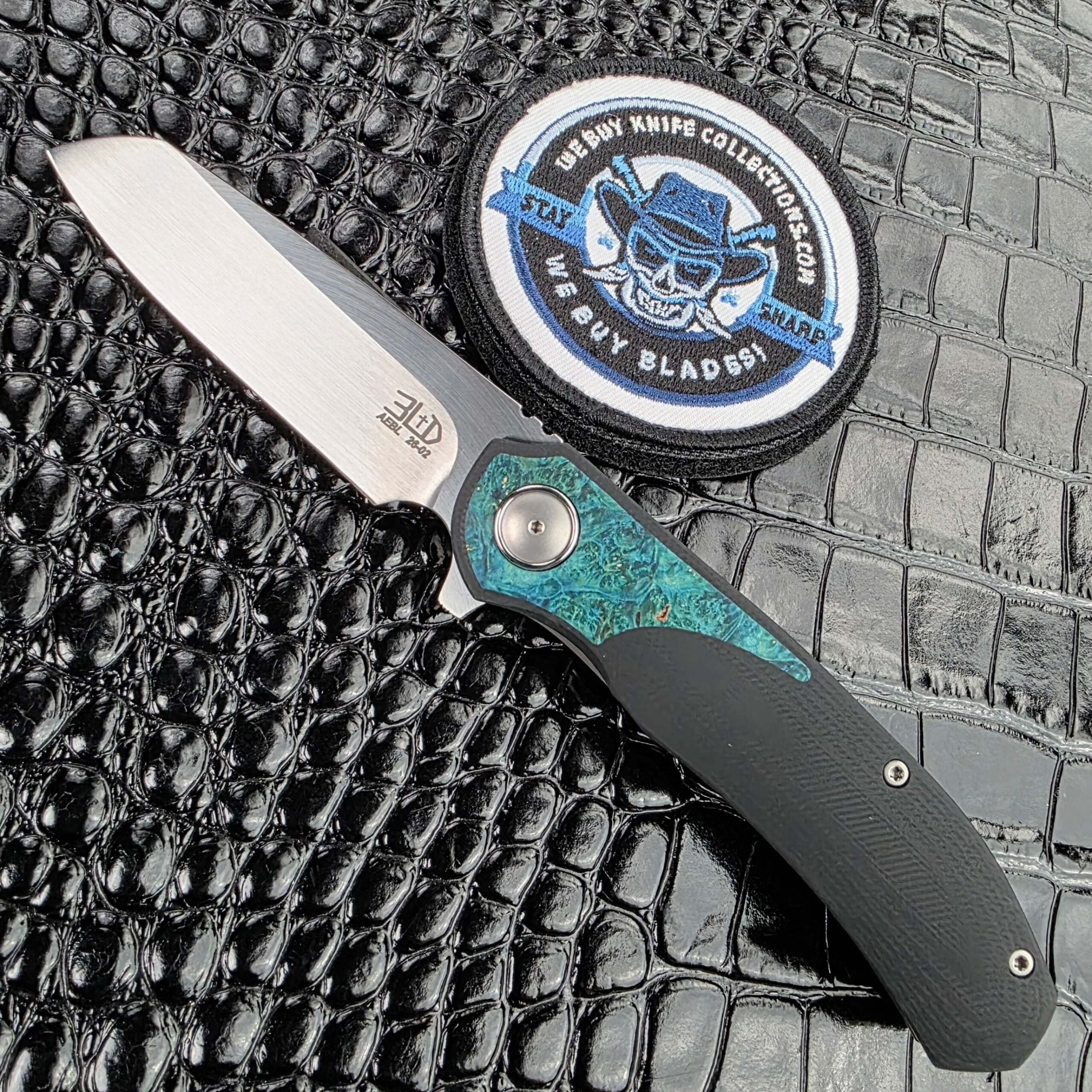 3LegDog M10 open on the show side, hand-rubbed satin AEB-L drop point above a teal stabilised burl inlay, with the We Buy Knife Collections patch in frame