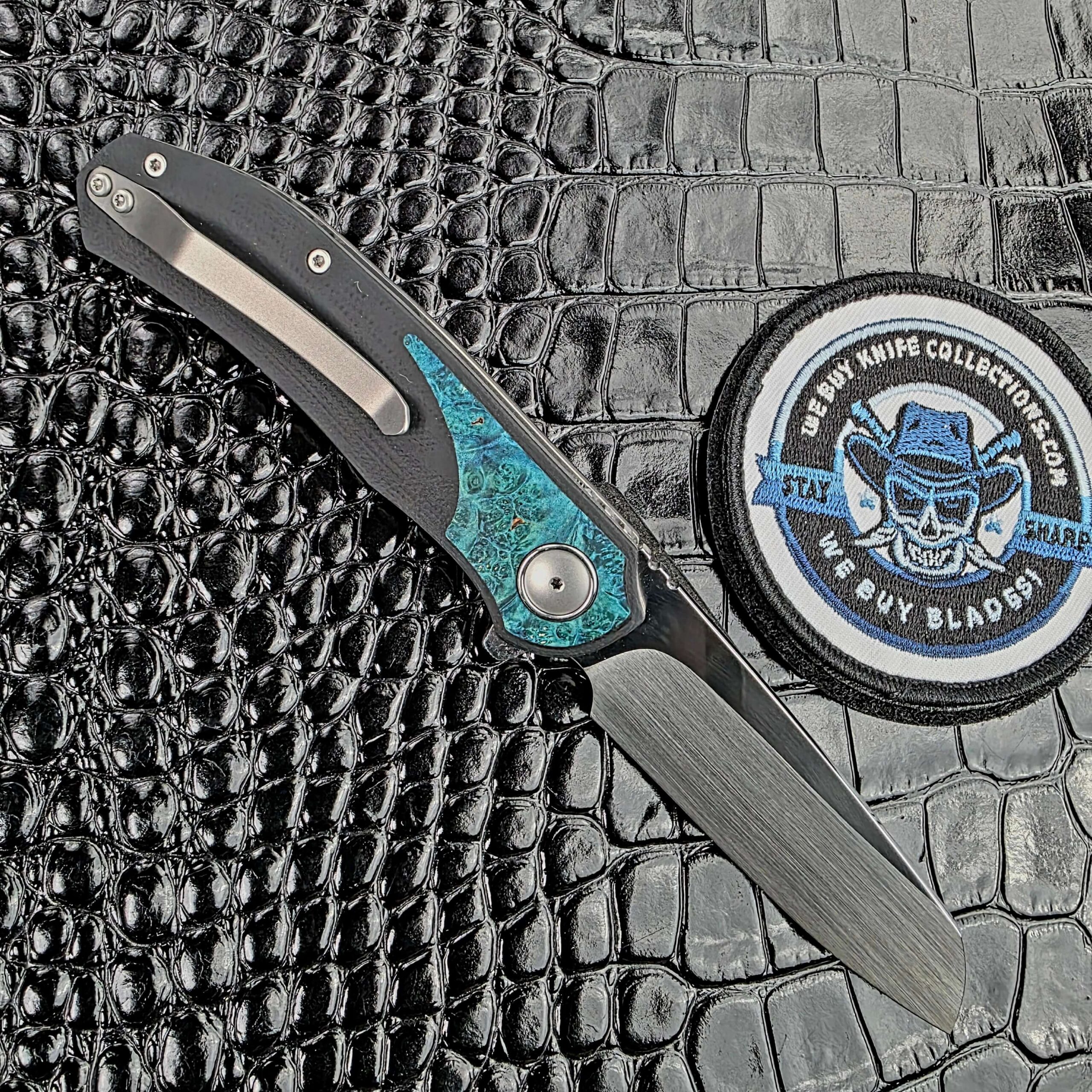 3LegDog M10 open on the clip side, showing the titanium pocket clip and teal burl inlay, with the We Buy Knife Collections patch in frame