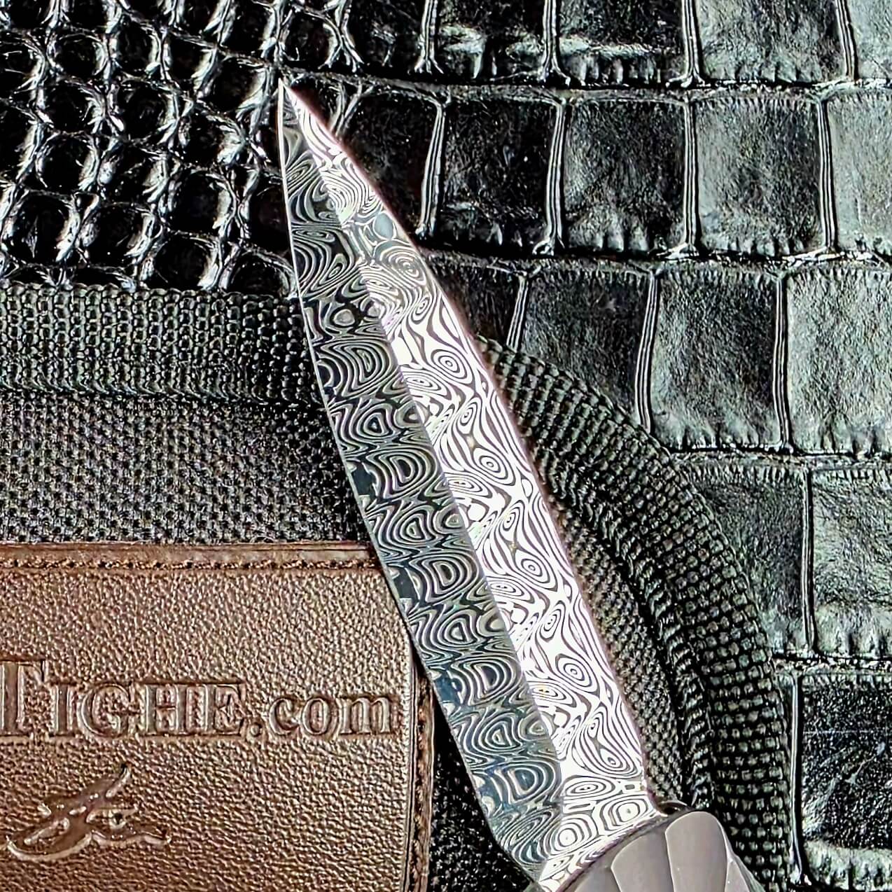 Damasteel dagger blade of the Brian Tighe Tighe Twist OTF beside the maker's branded soft case