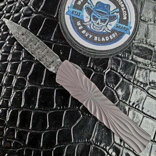 Brian Tighe Tighe Twist OTF deployed on the show side, Damasteel dagger blade against a DLC-coated fluted titanium handle, with the We Buy Knife Collections patch in frame