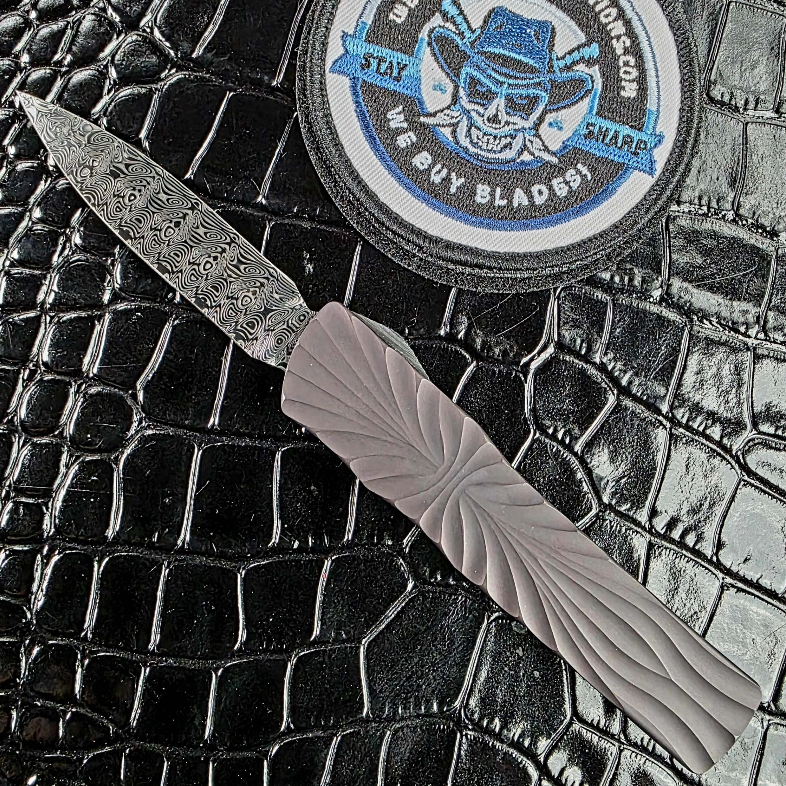 Brian Tighe Tighe Twist OTF deployed on the show side, Damasteel dagger blade against a DLC-coated fluted titanium handle, with the We Buy Knife Collections patch in frame