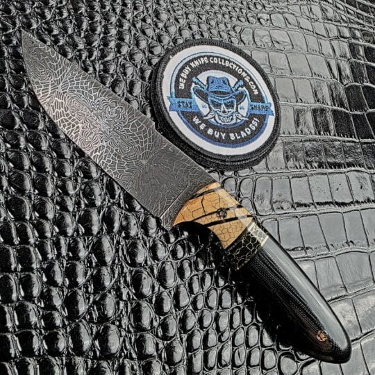Desert Sage Desert Dweller fixed blade on the show side, 2500 layer mosaic damascus above mammoth tusk bolsters and a sculpted black G10 handle, with the We Buy Knife Collections patch in frame