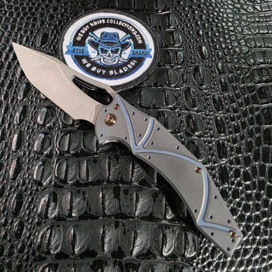 Ferrum Forge x Gavko Spinner open on the show side, stonewashed CPM 20CV blade above a Facets milled titanium handle multi-anodized blue, with the We Buy Knife Collections patch in frame