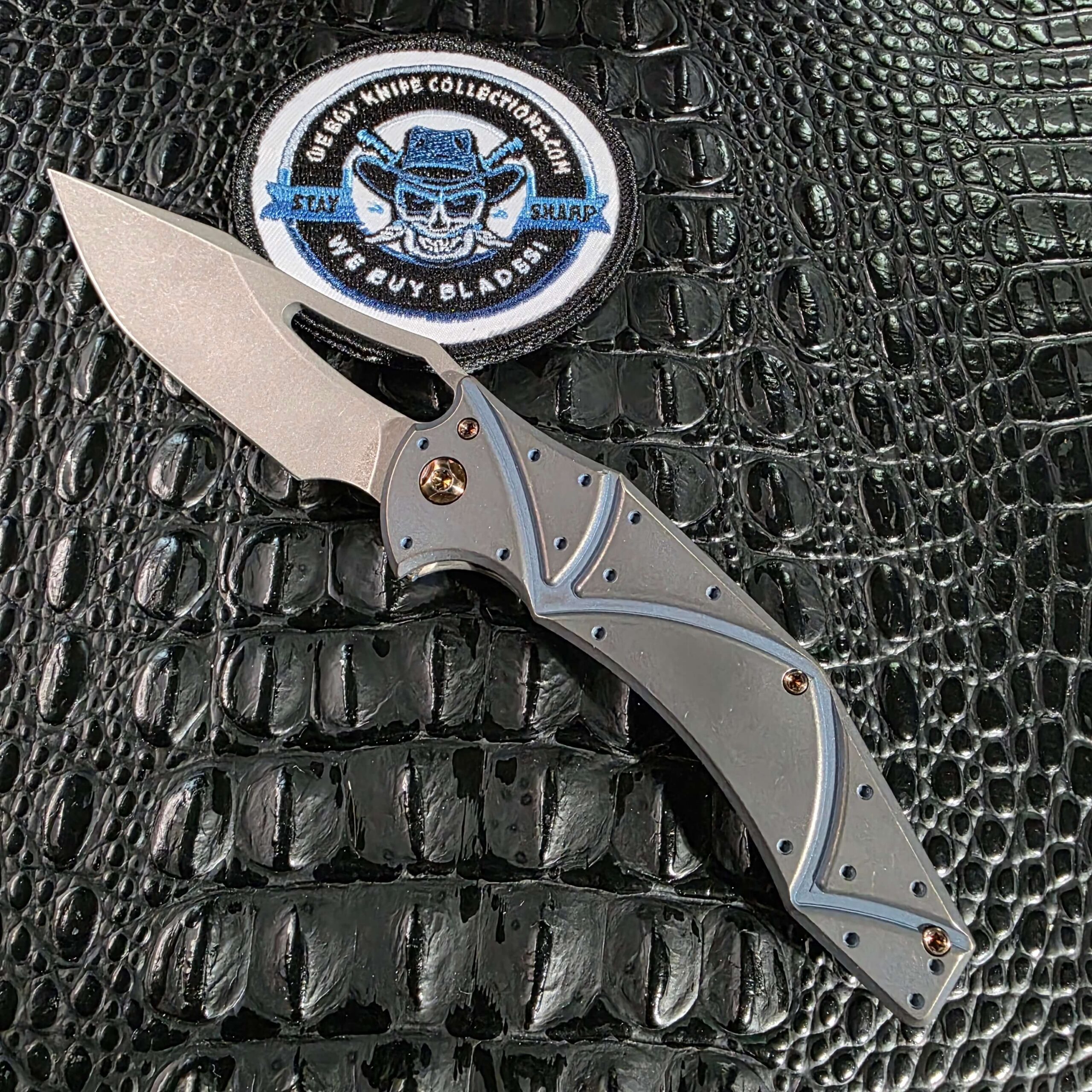 Ferrum Forge x Gavko Spinner open on the show side, stonewashed CPM 20CV blade above a Facets milled titanium handle multi-anodized blue, with the We Buy Knife Collections patch in frame