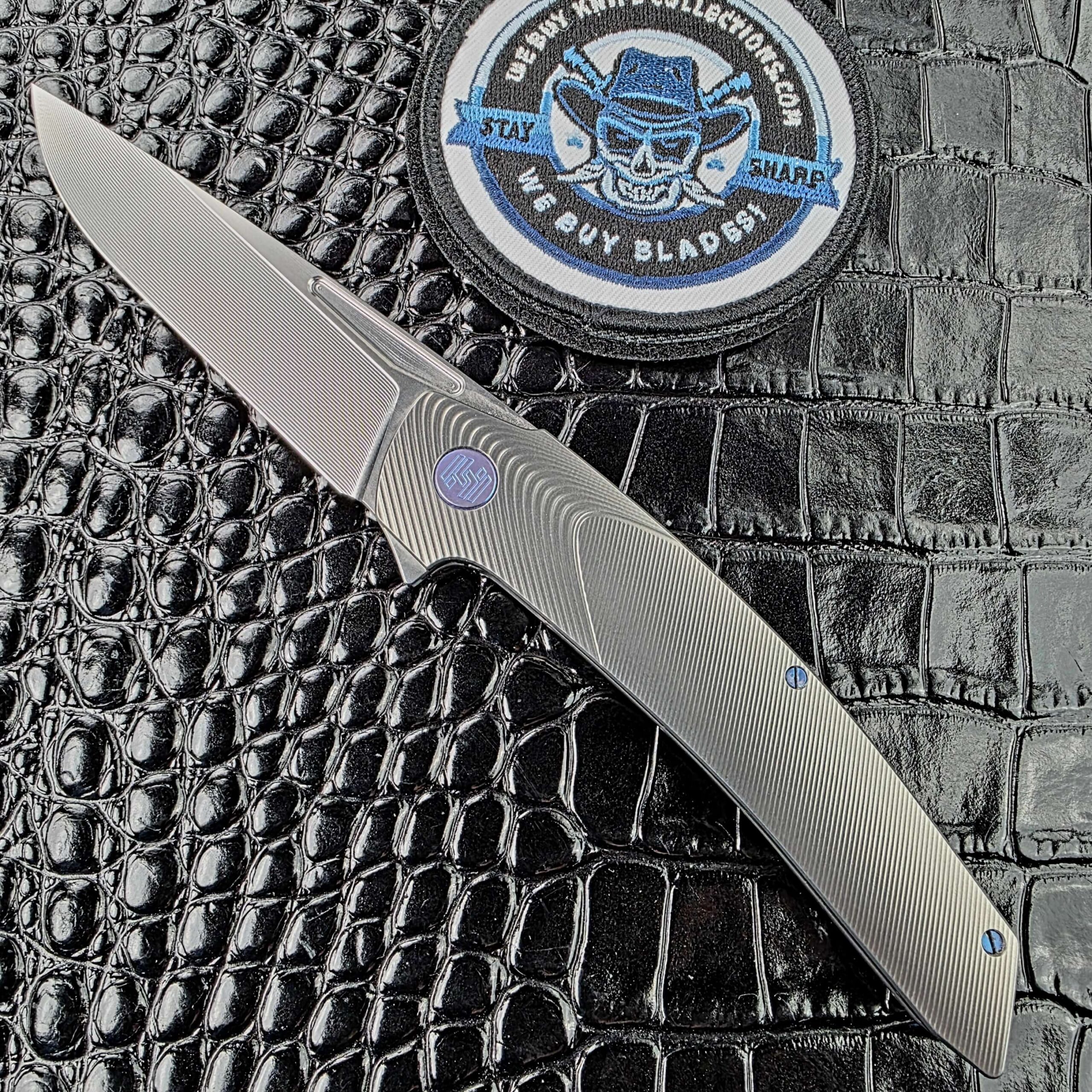 Hog House Model T Gen 2 open on the show side, RWL-34 satin blade with step-milled bevel above a Gunmetal titanium handle, with the We Buy Knife Collections patch in frame