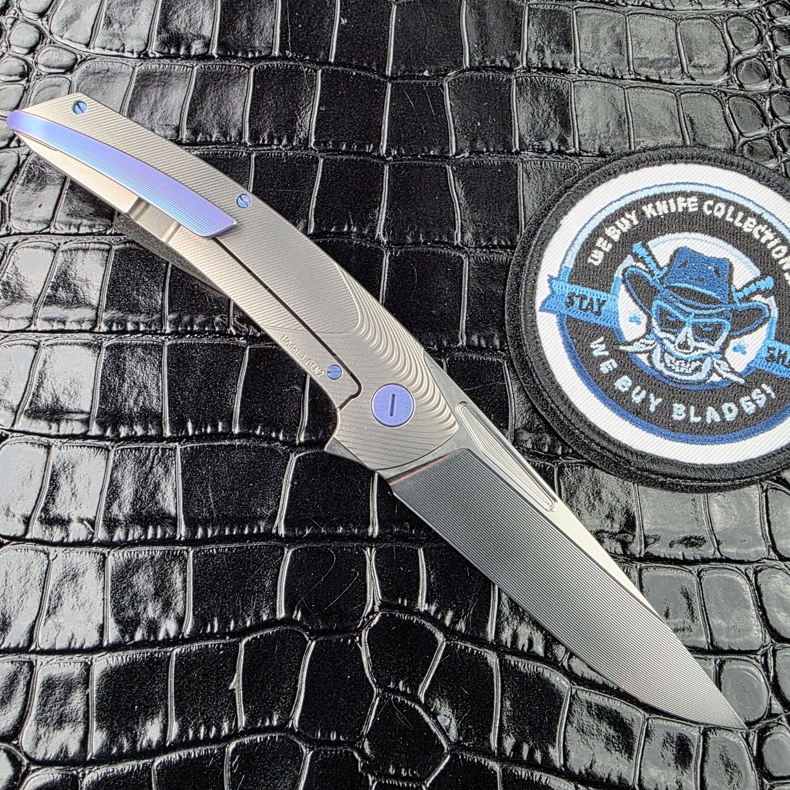 Hog House Model T Gen 2 open on the clip side showing the Blue Chameleon anodised titanium pocket clip, with the We Buy Knife Collections patch in frame