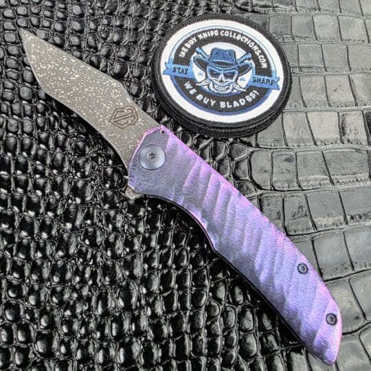 John Gray Slitter V2 Recurve open on the show side, Dark Rain MagnaCut recurve blade above a blurple sculpted titanium handle, with the We Buy Knife Collections patch in frame