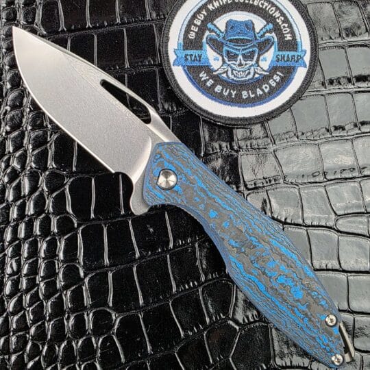 Koenig Arius Gen 4 open on the show side, CPM 20CV blade with polished flats above a marbled blue carbon fiber scale, with the We Buy Knife Collections patch in frame