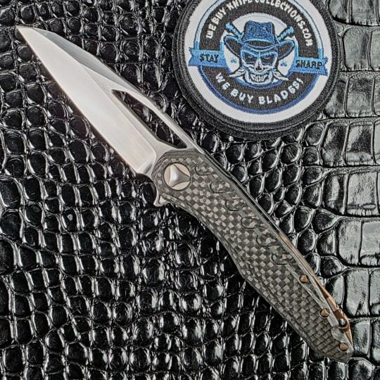 Marfione Custom Sigil open on the show side, hand-rubbed satin Elmax wharncliffe against a 3D milled carbon fiber frame, with the We Buy Knife Collections patch in frame