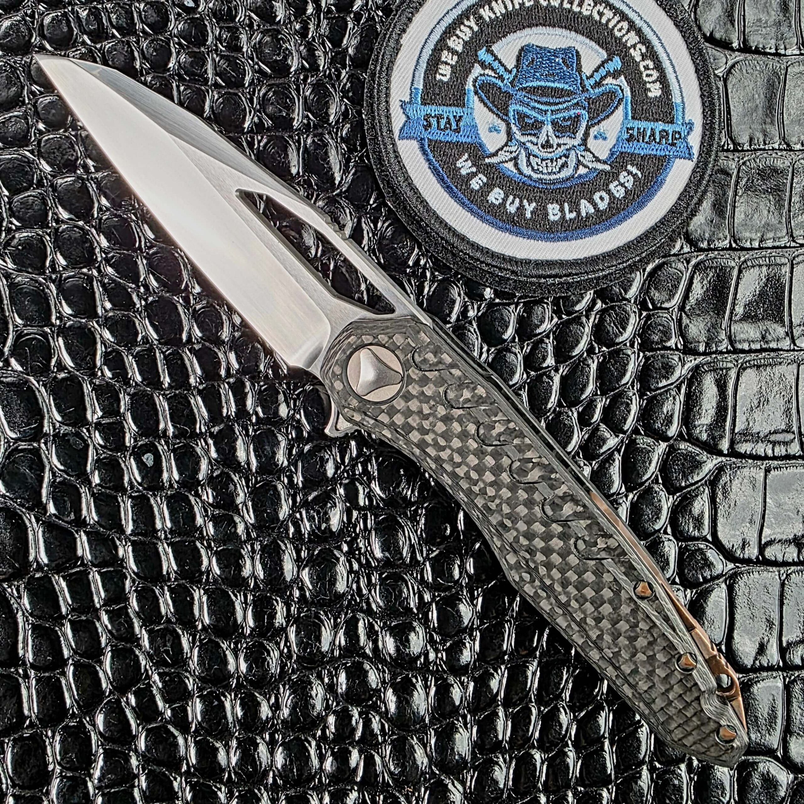 Marfione Custom Sigil open on the show side, hand-rubbed satin Elmax wharncliffe against a 3D milled carbon fiber frame, with the We Buy Knife Collections patch in frame