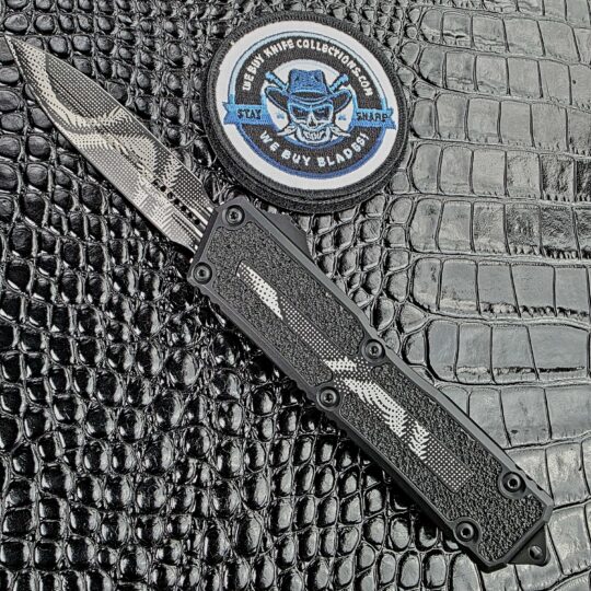 Microtech Scarab II Gen III deployed on the show side, Geo Trace Camo D/E dagger blade and aluminium handle with 3M grip inserts, with the We Buy Knife Collections patch in frame