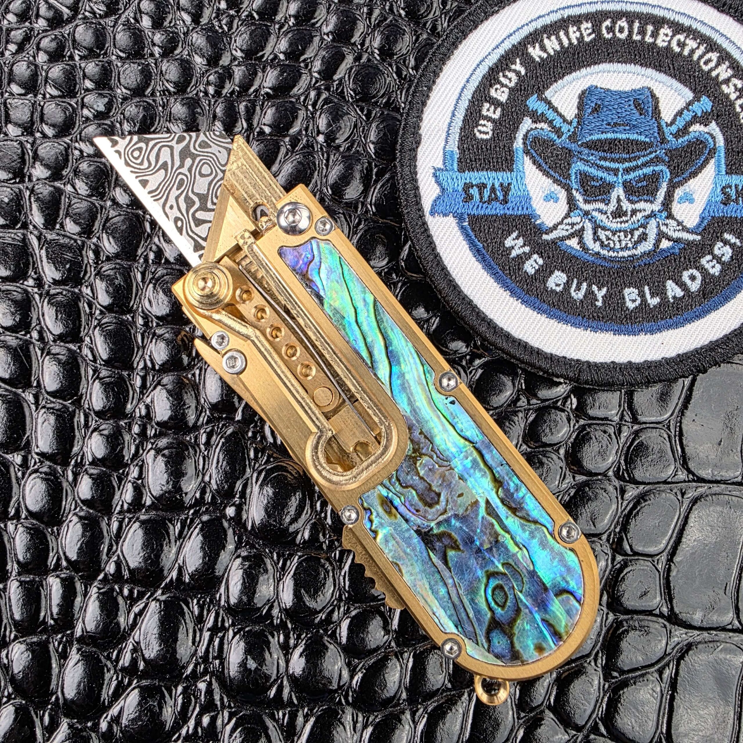 Miroc Razor Knife OTF Automatic Custom deployed on the show side, laser etched utility blade above a brass chassis with abalone inlay, with the We Buy Knife Collections patch in frame