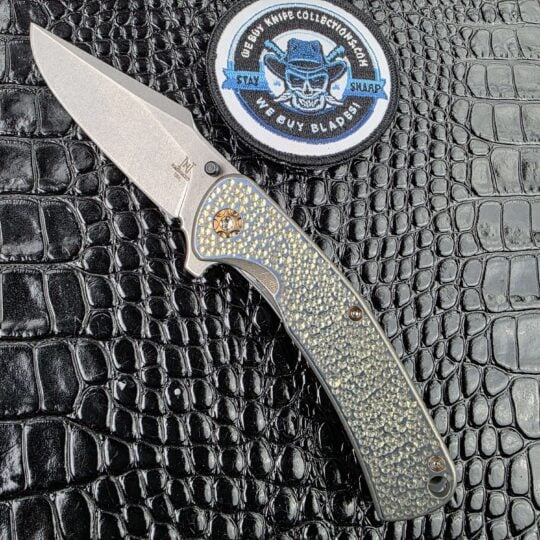 Nicholas Nichols Guppy 3.5 open on the show side, CPM MagnaCut drop point above a blue-to-bronze anodized Pebble Wave titanium handle, with the We Buy Knife Collections patch in frame