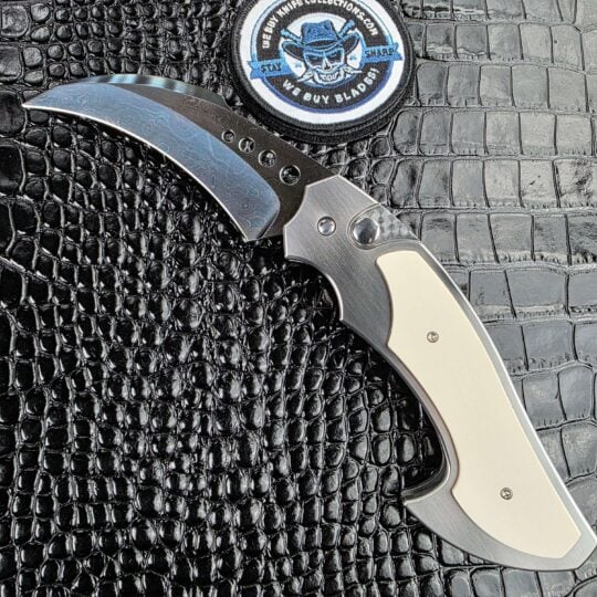 Reese Weiland Large Rastor open on the show side, Brad Vice blued damascus hawkbill above a stainless frame with white mammoth tusk scales, with the We Buy Knife Collections patch in frame
