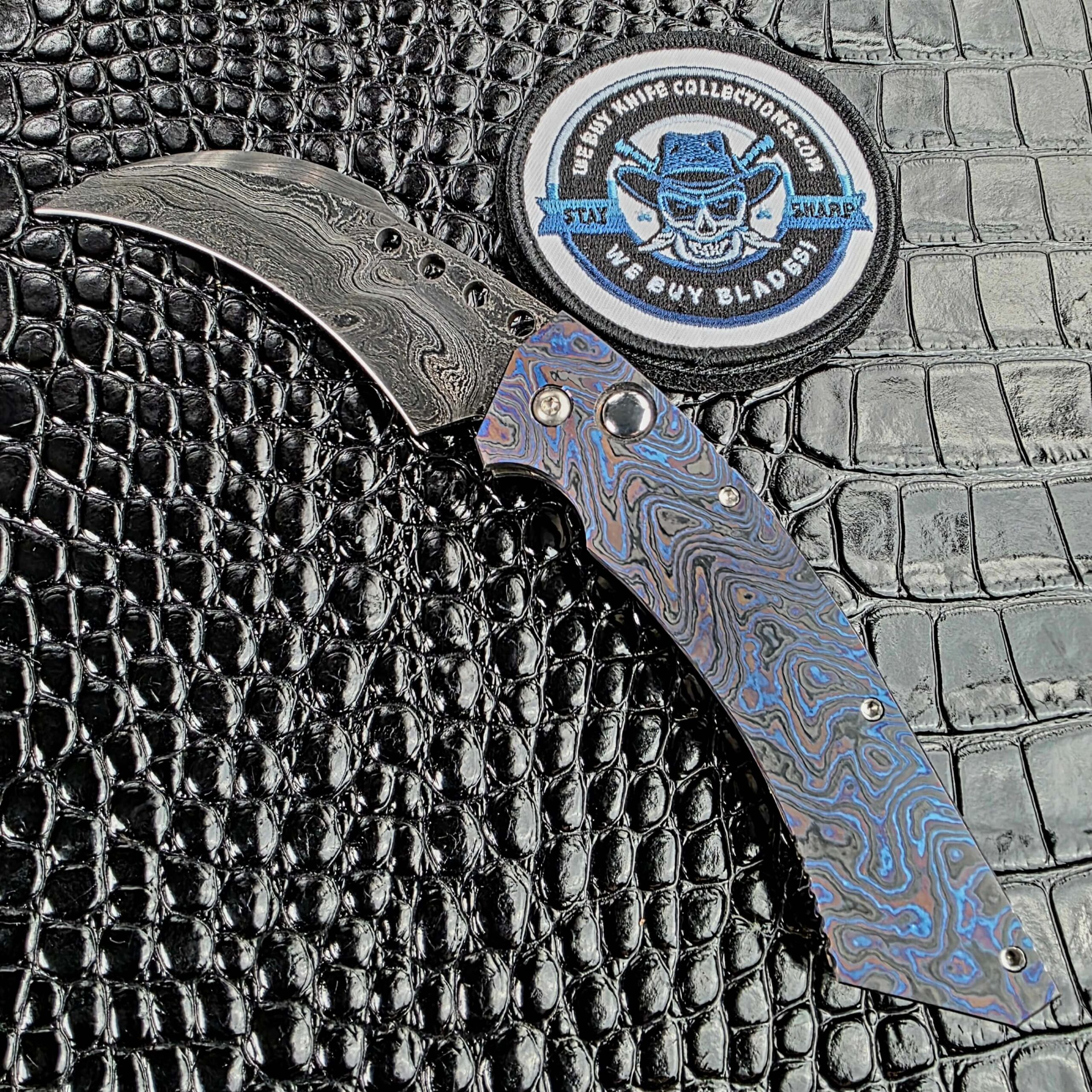 Reese Weiland Hawkbill 2nd Gen open on the show side, damascus blade against a full Timascus frame, with the We Buy Knife Collections patch in frame
