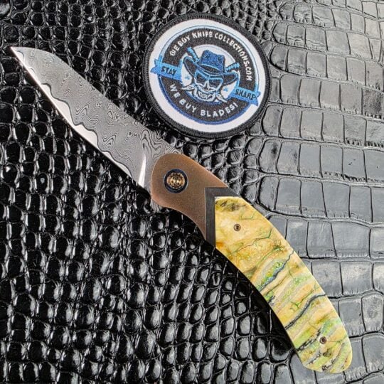 Andriy Mozil Model 22-26 Custom open on the show side, MagnaMax san mai Damascus blade above stabilized mammoth tooth scales, with the We Buy Knife Collections patch in frame