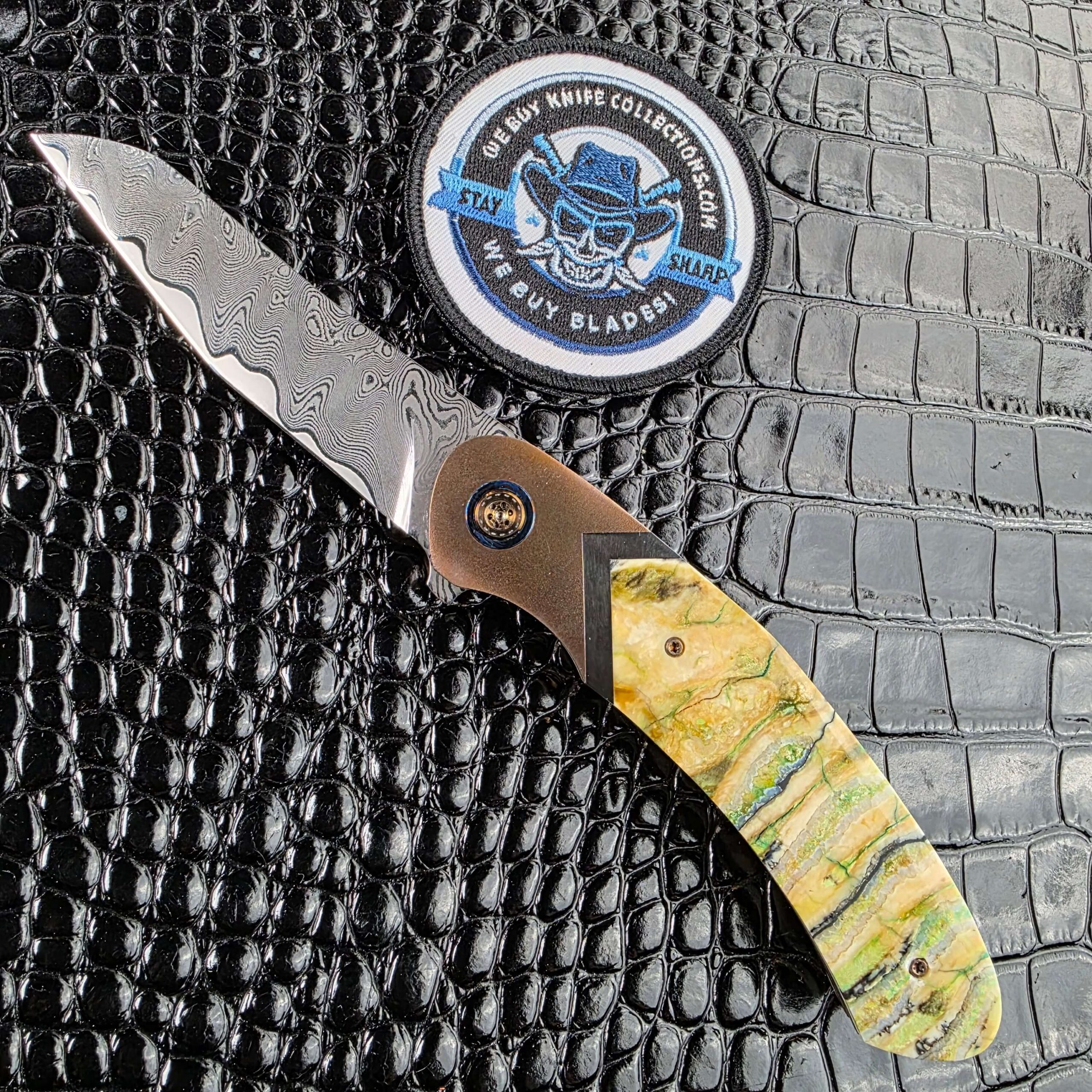Andriy Mozil Model 22-26 Custom open on the show side, MagnaMax san mai Damascus blade above stabilized mammoth tooth scales, with the We Buy Knife Collections patch in frame