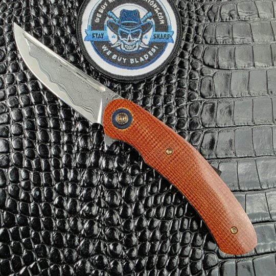 Andriy Mozil Model 23-26 open on the show side, Elmax san mai Damascus blade above canvas Micarta scales with a Timascus bolster, with the We Buy Knife Collections patch in frame