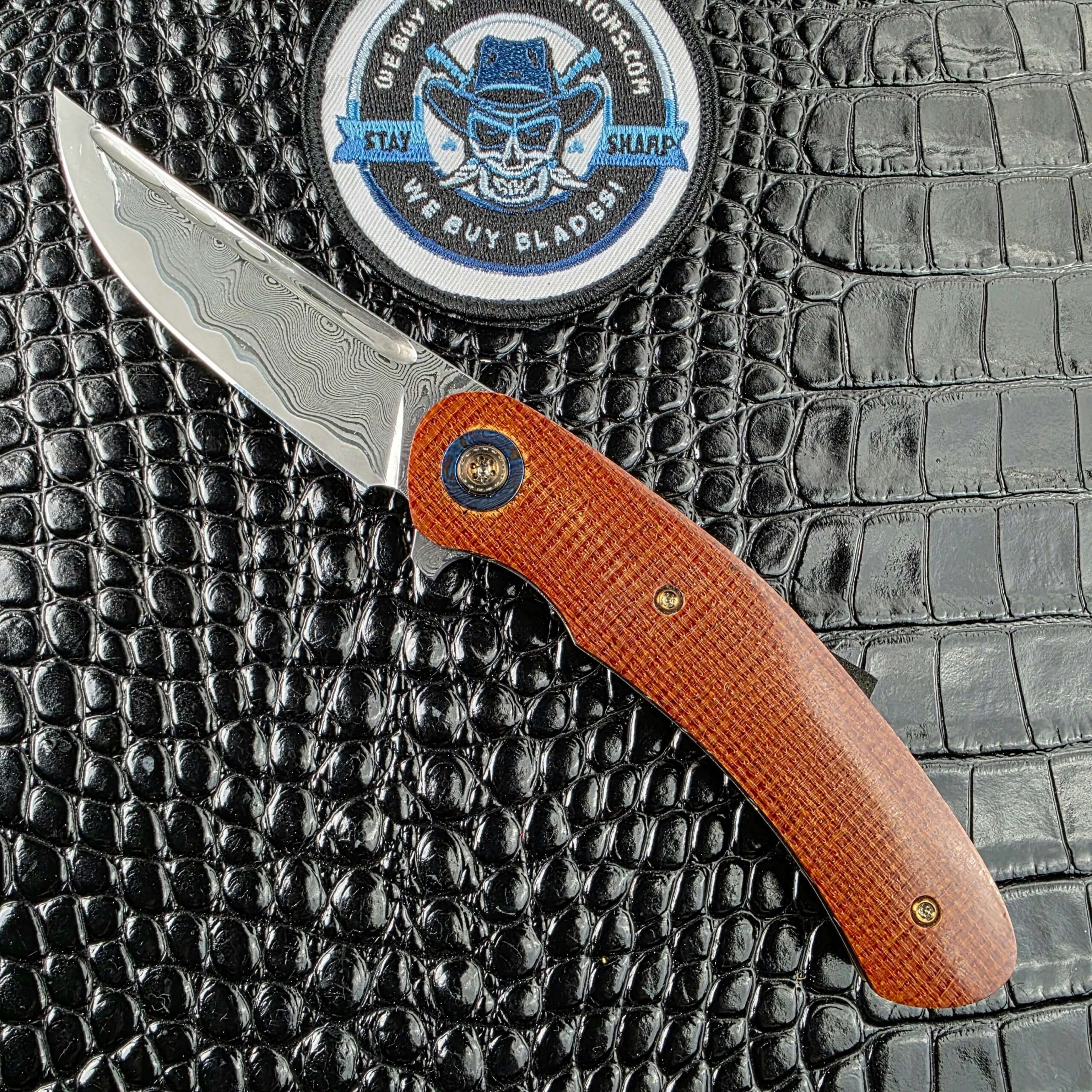 Andriy Mozil Model 23-26 open on the show side, Elmax san mai Damascus blade above canvas Micarta scales with a Timascus bolster, with the We Buy Knife Collections patch in frame