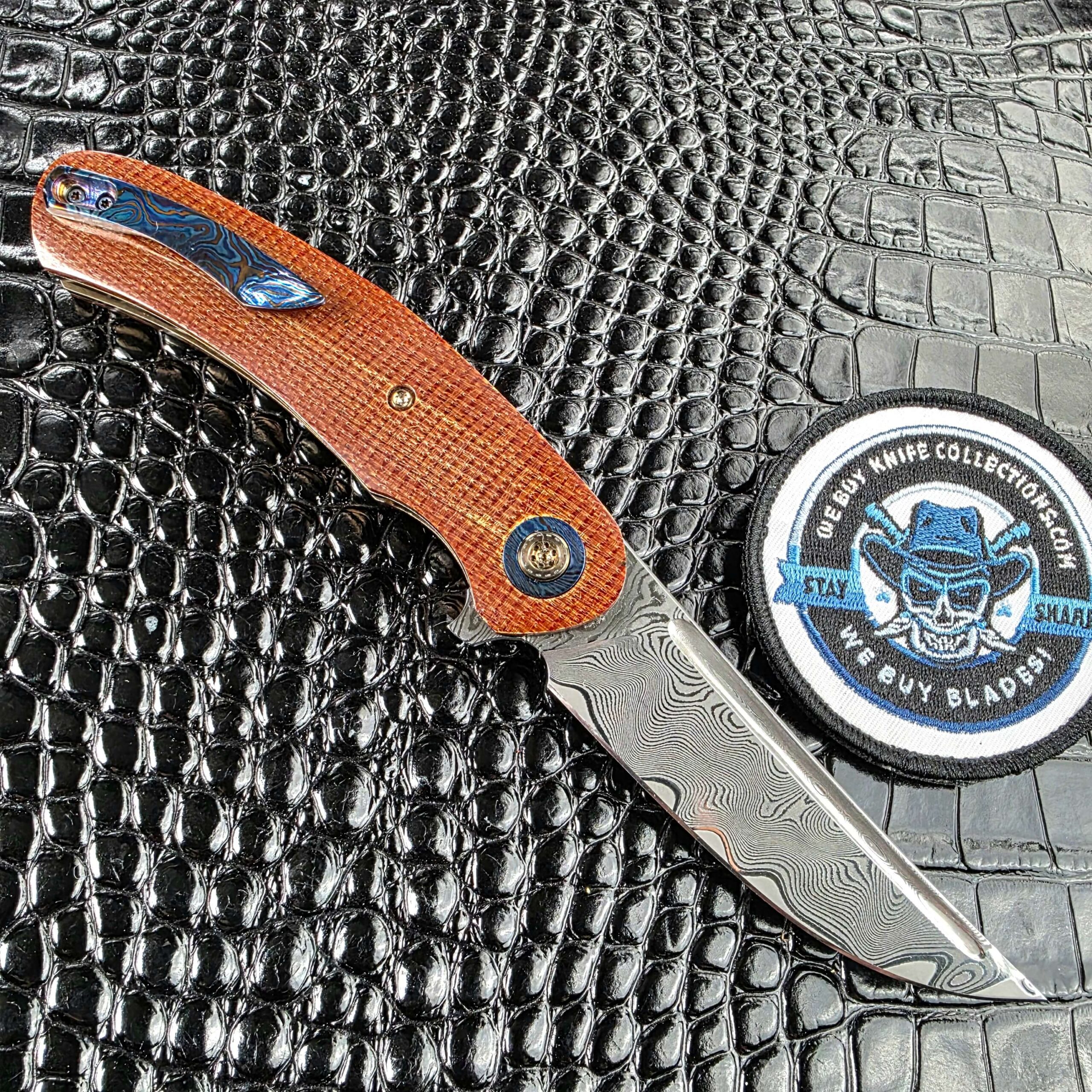 Andriy Mozil Model 23-26 open on the clip side showing the Timascus pocket clip, with the We Buy Knife Collections patch in frame