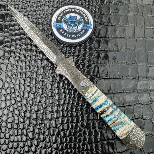 Burn Knives Automatic Dagger open on the show side, Chad Nichols Damascus dagger blade above mammoth molar scales, with the We Buy Knife Collections patch in frame