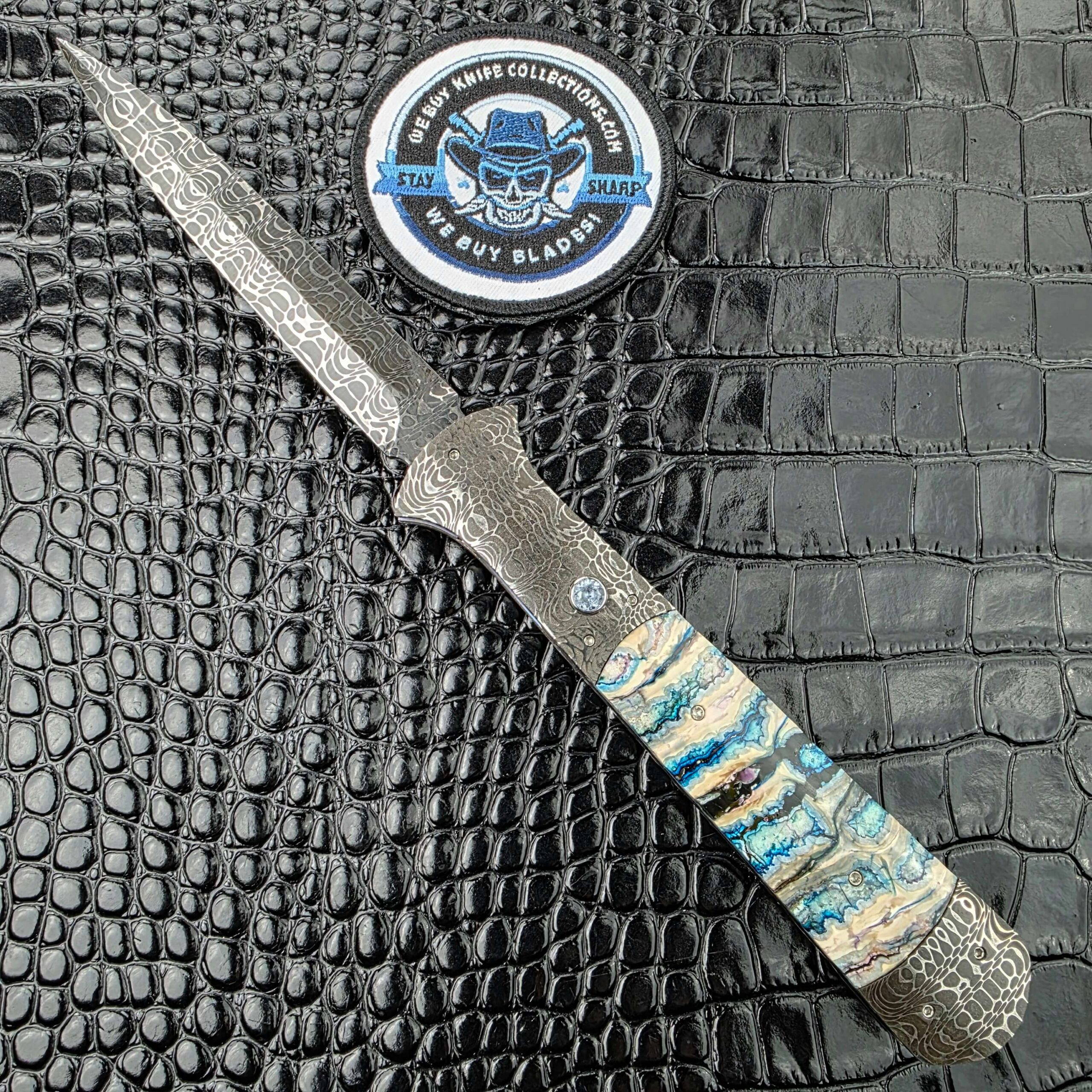 Burn Knives Automatic Dagger open on the show side, Chad Nichols Damascus dagger blade above mammoth molar scales, with the We Buy Knife Collections patch in frame
