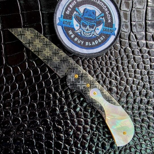 Curt Zimmermann Ranch Hand custom liner lock open on black crocodile leather, Mother of Pearl inlay and Mosaic Damascus frame beside the We Buy Knife Collections patch