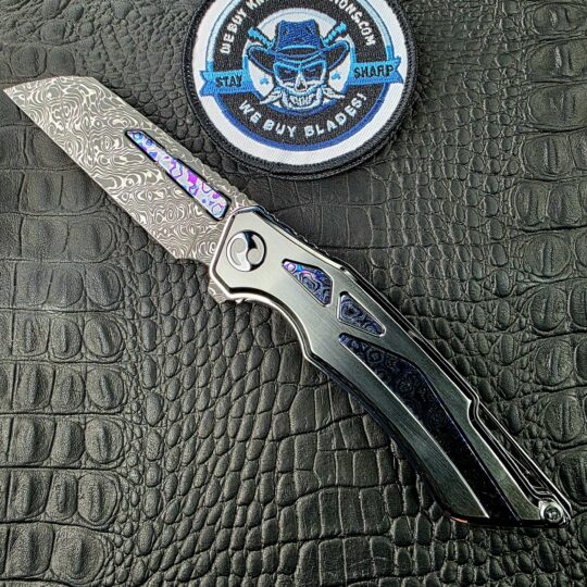 Dew Hara Seismic Damascus Ray Custom 1 of 1 open on its show side beside the We Buy Knife Collections patch