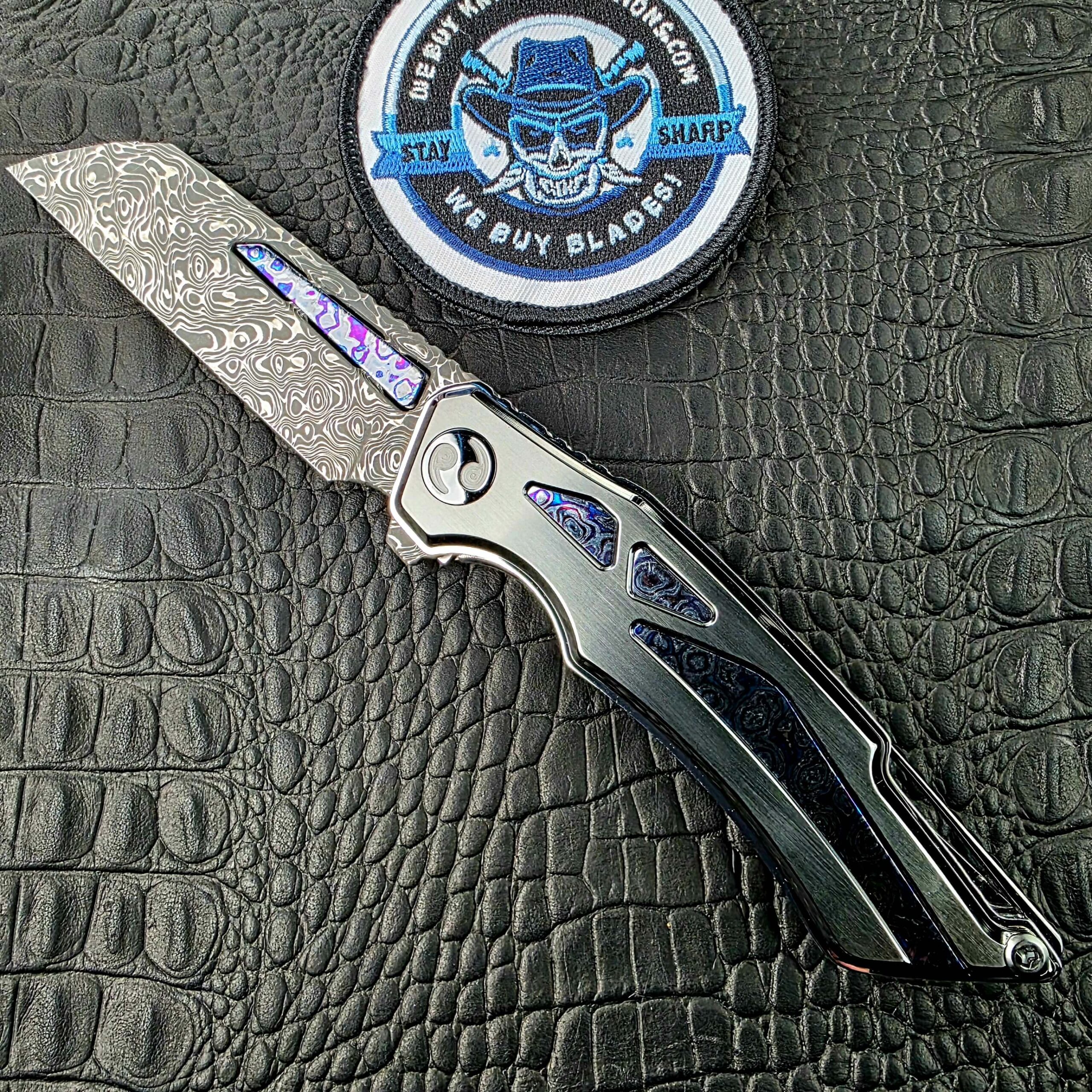 Dew Hara Seismic Damascus Ray Custom 1 of 1 open on its show side beside the We Buy Knife Collections patch