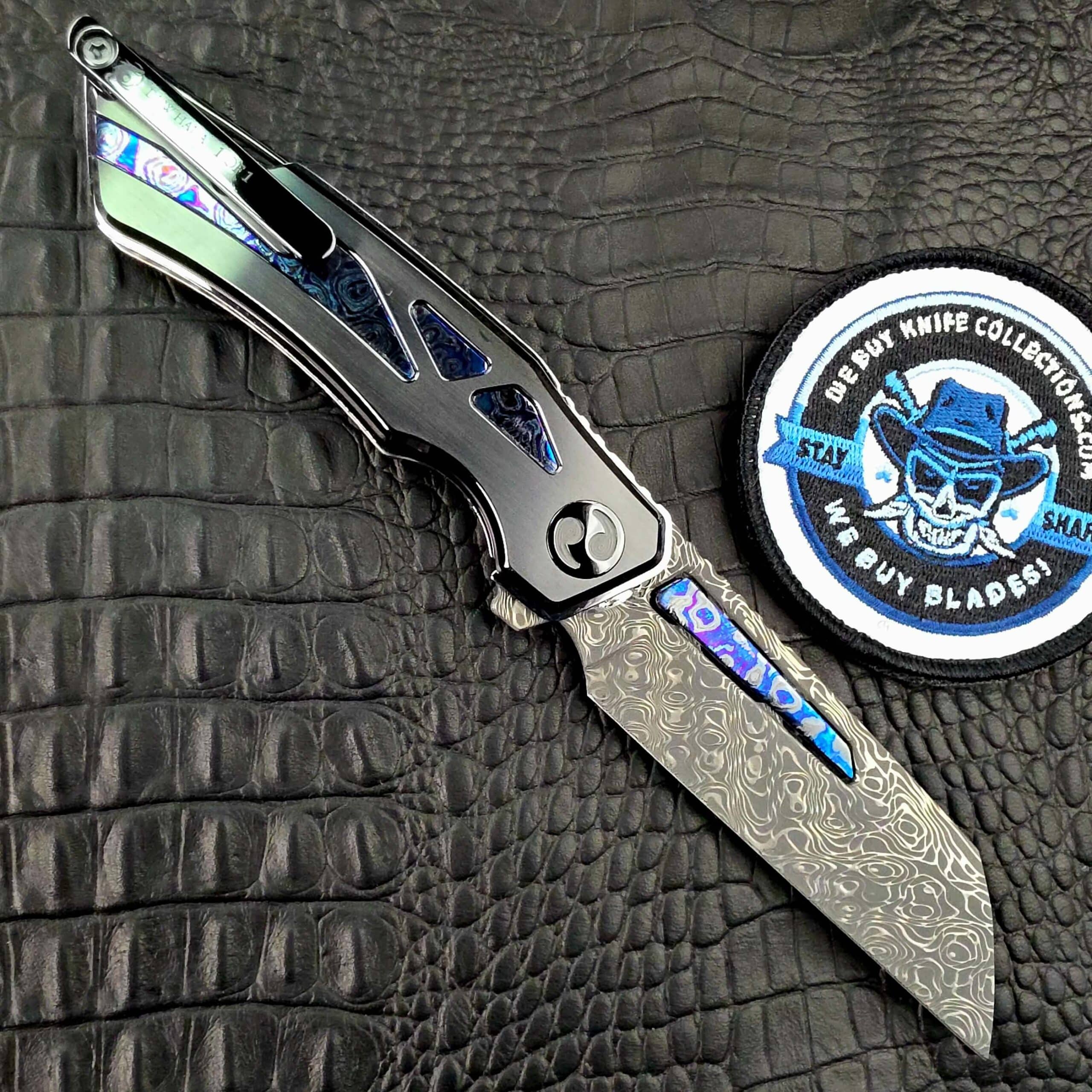 Dew Hara Seismic Damascus Ray open on its clip side with the DLC Titanium pocket clip and the We Buy Knife Collections patch