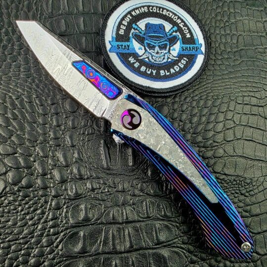 Dew Hara Sharktooth Damascus METEOR Custom 1 of 1 open on its show side beside the We Buy Knife Collections patch