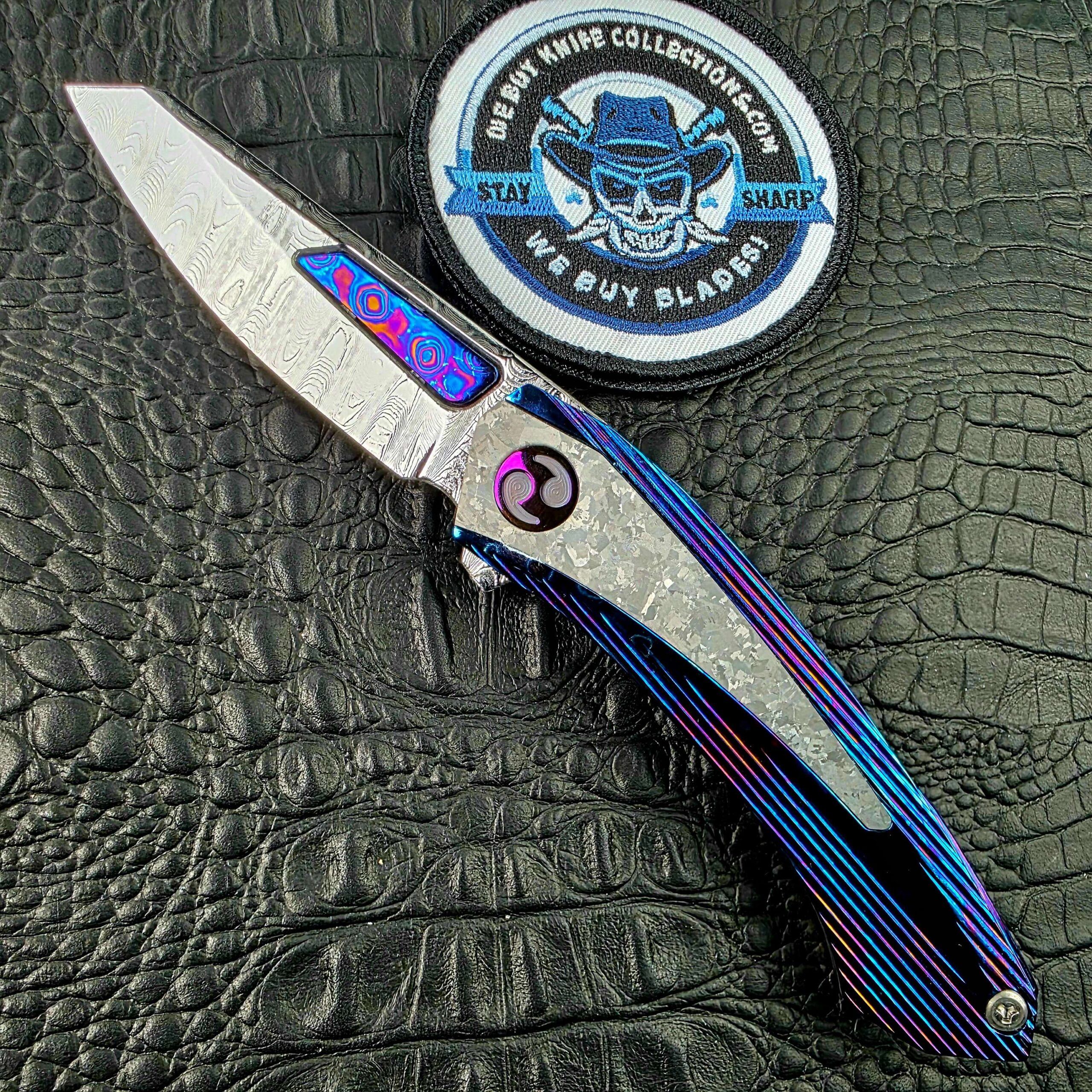 Dew Hara Sharktooth Damascus METEOR Custom 1 of 1 open on its show side beside the We Buy Knife Collections patch
