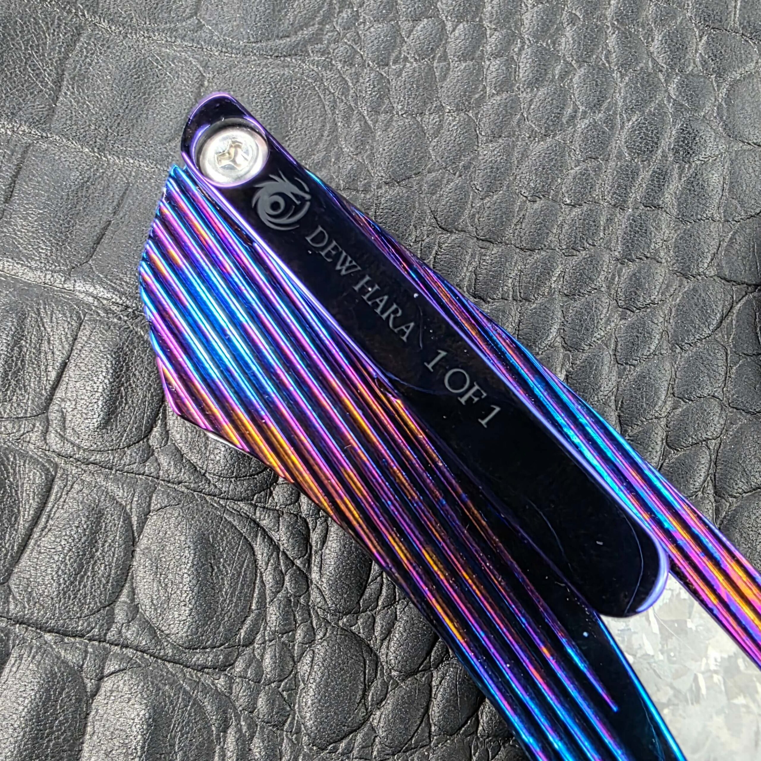 Titanium pocket clip on the Dew Hara METEOR marked with the Dew Hara logo and DEW HARA 1 OF 1, over grooved Timascus