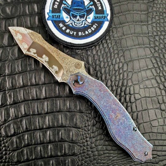 Glenn Waters Titan open on its show side with a Mitsudomoe pivot and multi-color tungsten carbide frame beside the We Buy Knife Collections patch